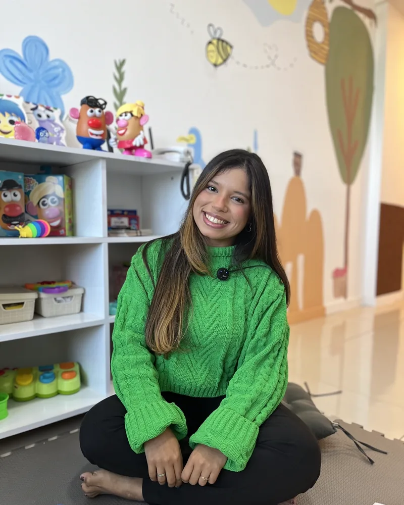Jessica Costa, psicóloga infantojuvenil, sentada na sala lúdica do consultório com sorriso aberto, blusa verde, brinquedos coloridos e mural de abelhinha ao fundo
