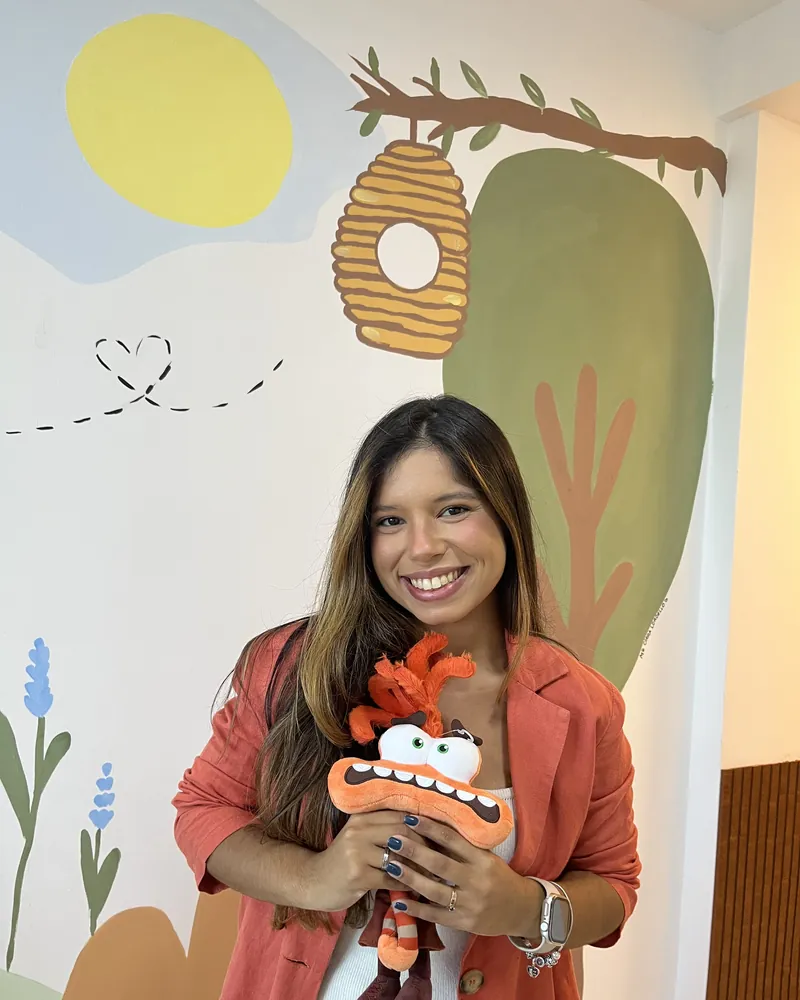 Jessica Costa Psi, psicóloga infantojuvenil, sorrindo em seu consultório com brinquedo terapêutico em mãos e mural infantil ao fundo